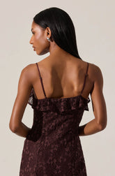 A woman is wearing a sleeveless, burgundy dress with thin spaghetti straps. The dress features a textured lace overlay with floral patterns and a ruffle detail along the top edge. The back of the dress is fitted, highlighting a sleek silhouette.